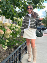 Load image into Gallery viewer, Woman in a striped cardigan and white skirt standing on a sidewalk with greenery and cars in the background.