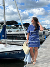 Load image into Gallery viewer, Woman in a blue dress standing on a dock next to a sailboat with a cloudy sky in the background.