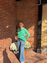 Load image into Gallery viewer, Woman in a green shirt and blue jeans standing against a brick wall with potted plants.