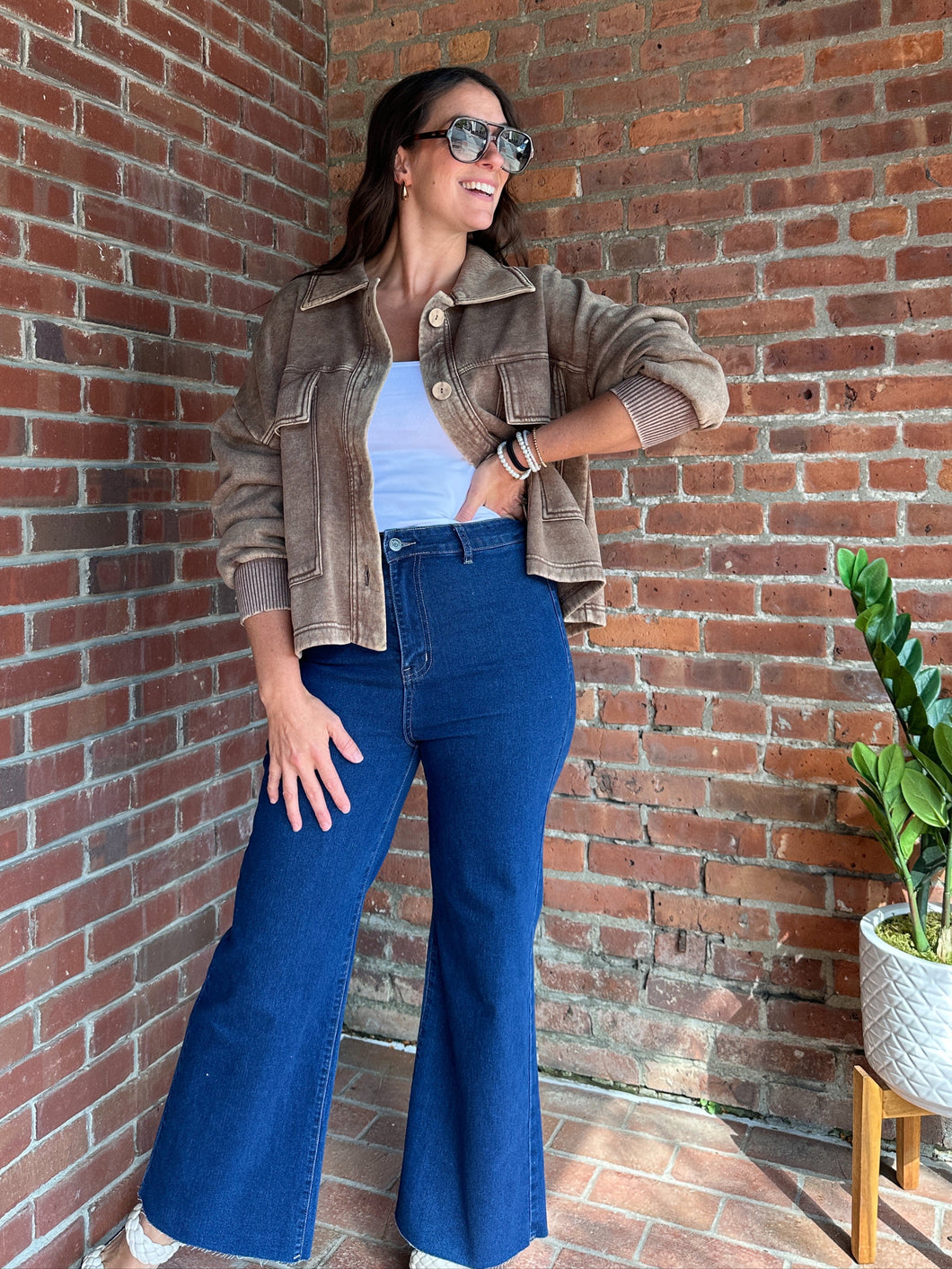 Woman in a brown jacket and blue jeans standing against a brick wall.