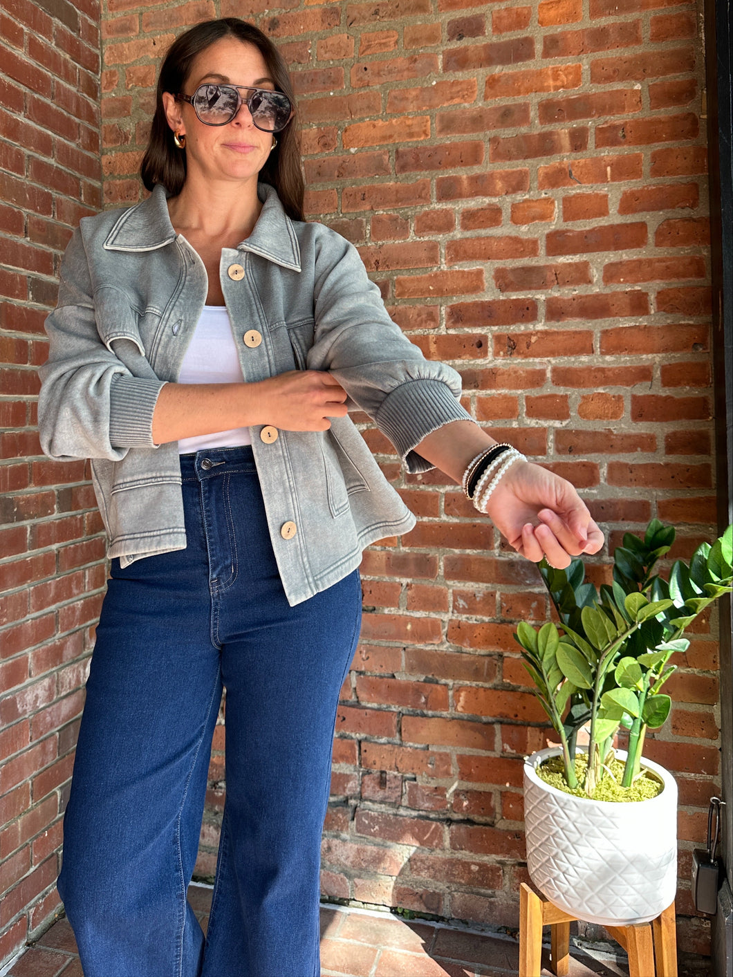 Woman in denim jacket and jeans standing against a brick wall with a plant nearby.