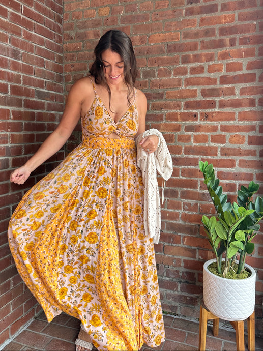 Woman in a yellow floral dress standing against a brick wall with a plant nearby.