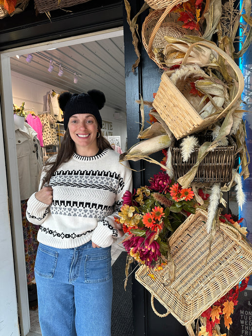 Black and cream fair isle sweater with black hat and oversized sticking on neck 

Person wearing a patterned sweater and jeans standing in front of a decorative display with corn stalks and flowers.