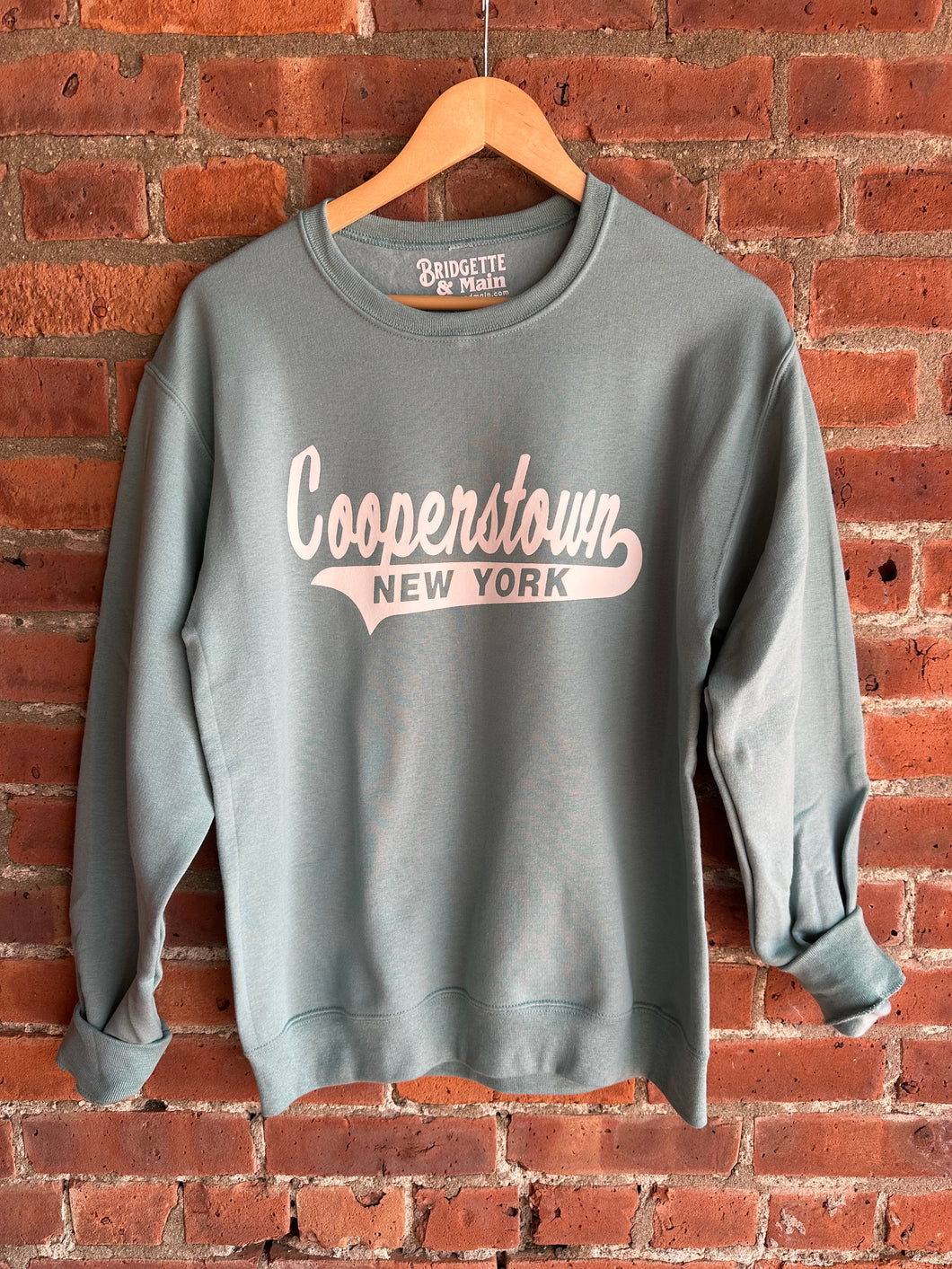 Sage Green sweatshirt with 'Cooperstown New York' text on a wooden hanger against a brick wall.