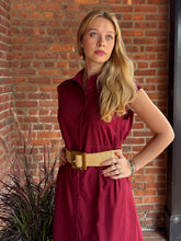 Load image into Gallery viewer, A woman with blonde hair wearing a sleeveless burgundy button-down dress with a wide tan woven belt, standing in front of a red brick wall.
